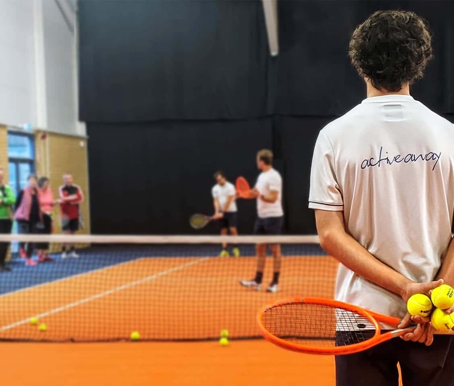 Tennis coach with a racket and balls watches players practising at David Lloyd Edinburgh indoor court.