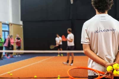 Tennis coach with a racket and balls watches players practising at David Lloyd Edinburgh indoor court.