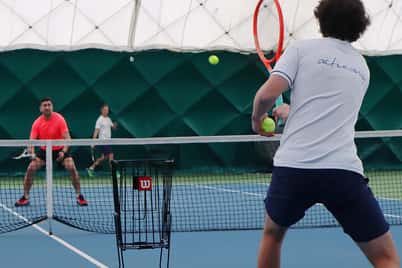 Tennis clinic at David Lloyd Northwood with two players engaged in an active rally on an indoor court under a dome.
