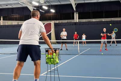 Group of tennis players in an indoor court at David Lloyd Southampton, with a coach instructing players near the net during a training drill.