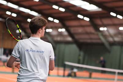 Tennis player in a clinic at David Lloyd West Bridgford's indoor tennis court, with tennis balls visible on the court.