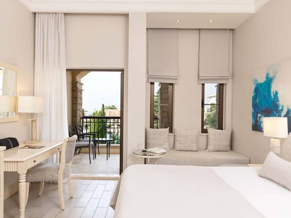 Deluxe double hotel room with neutral decor, a sitting area, and balcony overlooking a garden or pool.
