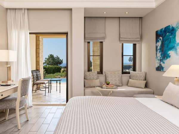 Luxurious hotel room with a double bed, lounge area, and desk, offering private terrace access with pool, golf, and sea views.