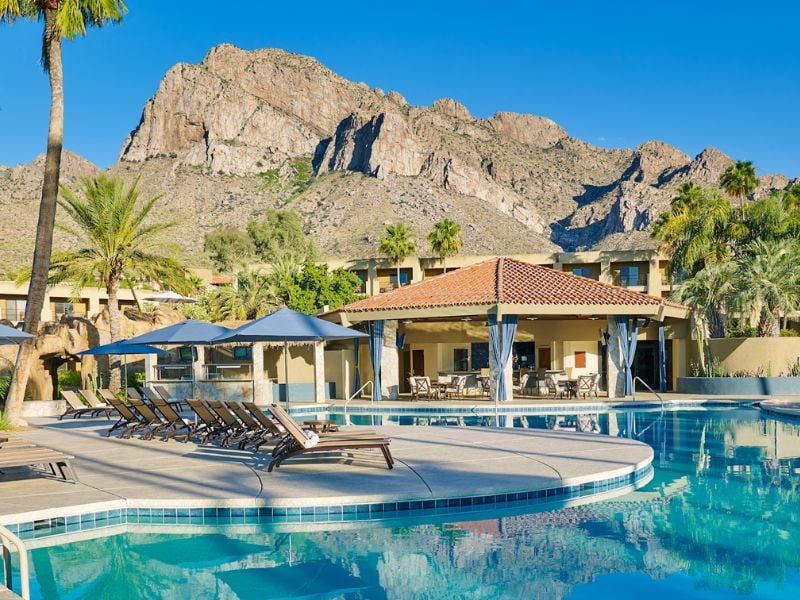Pool area at El Conquistador Resort with lounge chairs, parasols, and mountainous backdrop under a clear sky.
