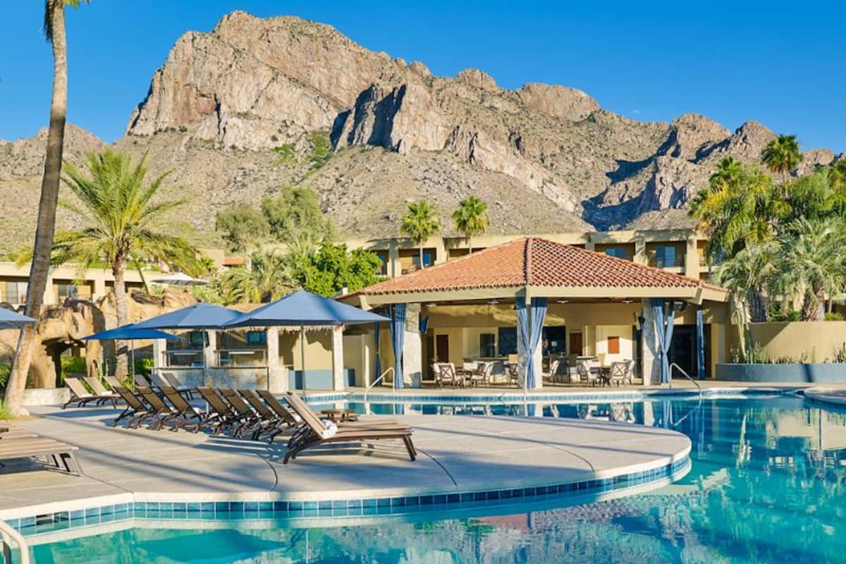 Pool area at El Conquistador Resort with lounge chairs, parasols, and mountainous backdrop under a clear sky.