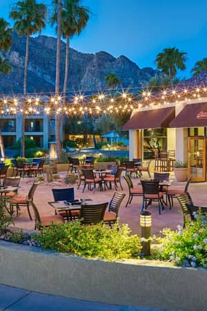 Outdoor dining area at El Conquistador Gallery with wooden tables, string lights, greenery, palm trees, and a mountainous backdrop.