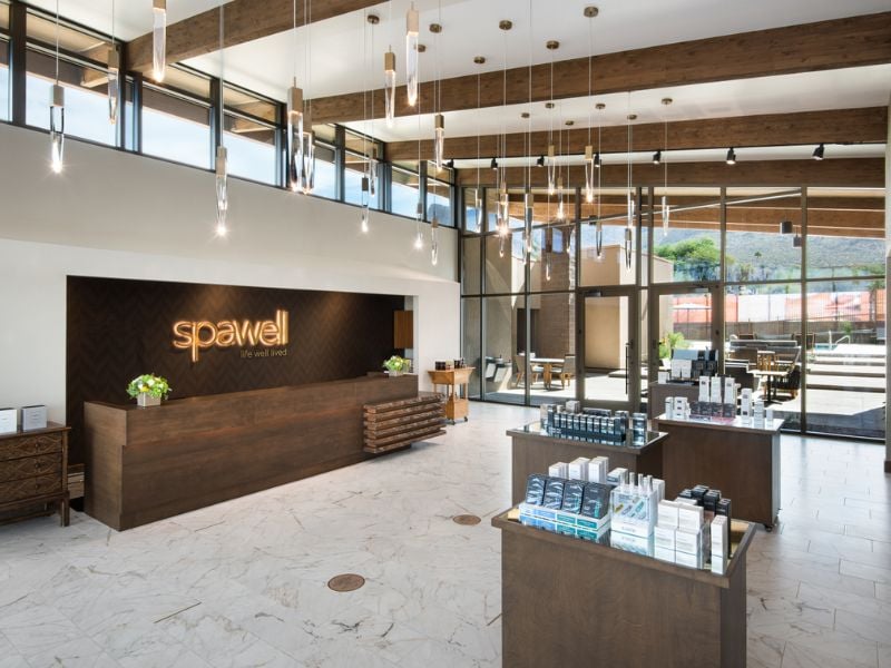 Luxurious spa interior at El Conquistador, featuring a wooden reception counter with 'spawell' logo, hanging lights, glass windows, and marble floors.