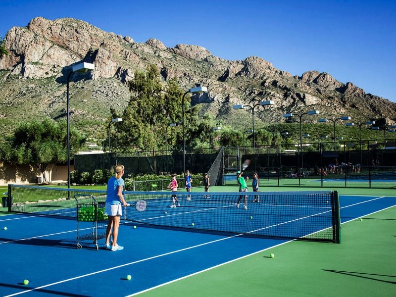 Outdoor tennis match at El Conquistador with players and coach, set against a backdrop of mountains and blue sky.