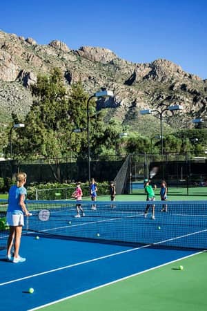 Outdoor tennis match at El Conquistador with players and coach, set against a backdrop of mountains and blue sky.