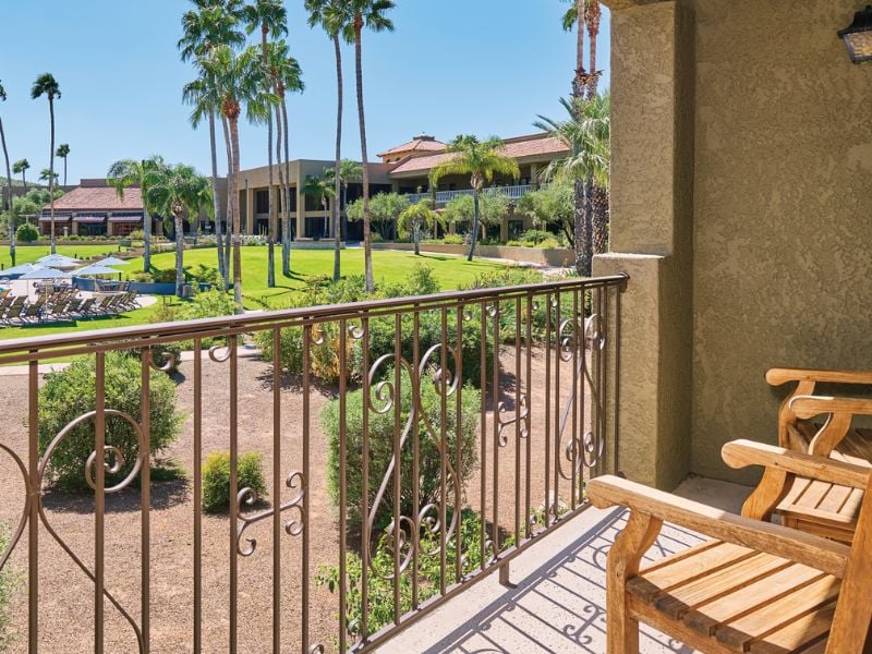 View from El Conquistador veranda with wooden chairs, overlooking a garden with palm trees and resort buildings.