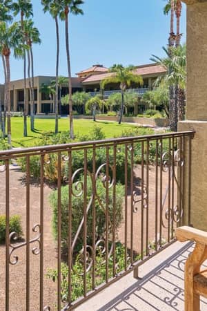 View from El Conquistador veranda with wooden chairs, overlooking a garden with palm trees and resort buildings.