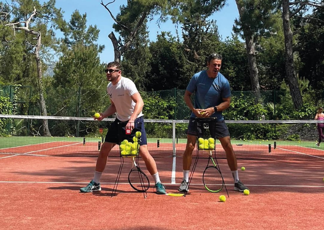 Two men balancing tennis ball hoppers on a red clay court surrounded by green trees, enjoying a playful moment.