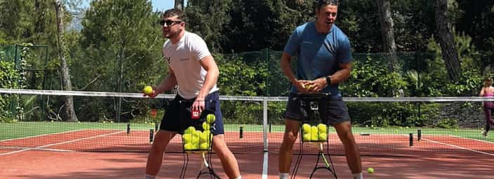 Two men balancing tennis ball hoppers on a red clay court surrounded by green trees, enjoying a playful moment.
