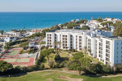 Aerial view of a seaside resort with apartments, a tennis court, and a golf course by the ocean.