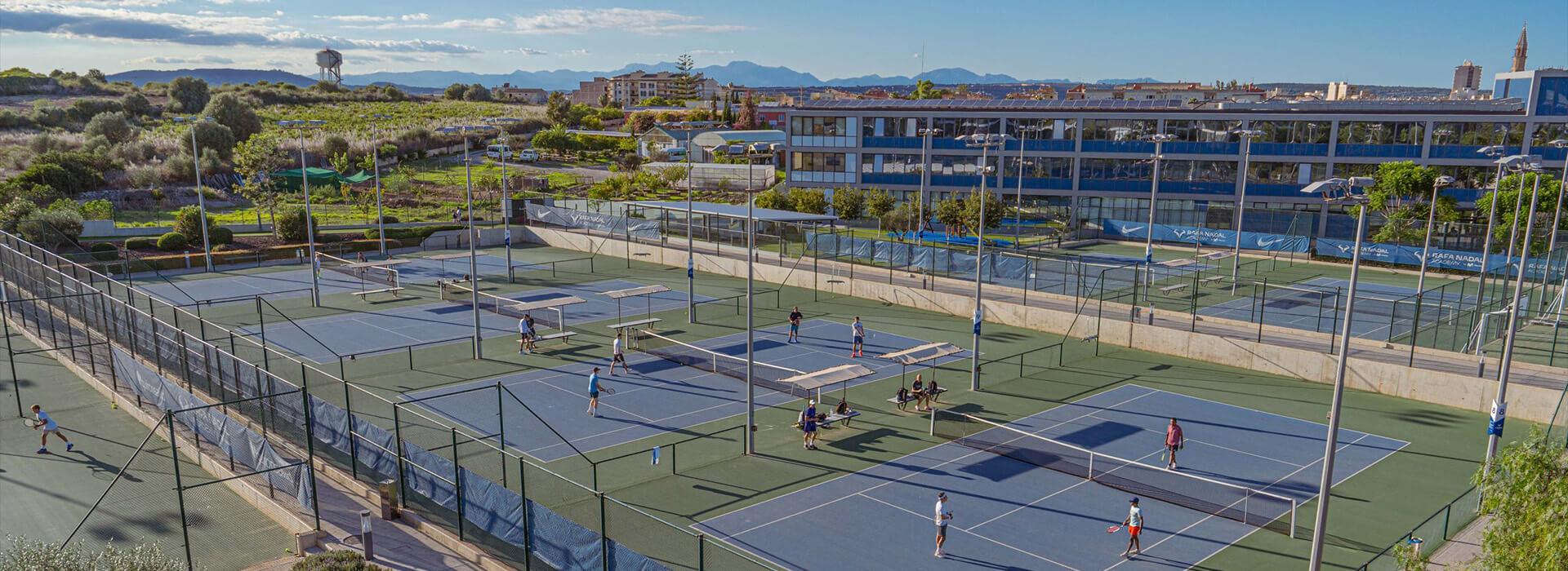 Overview of tennis academy with several courts, players practicing, and a modern building surrounded by greenery.