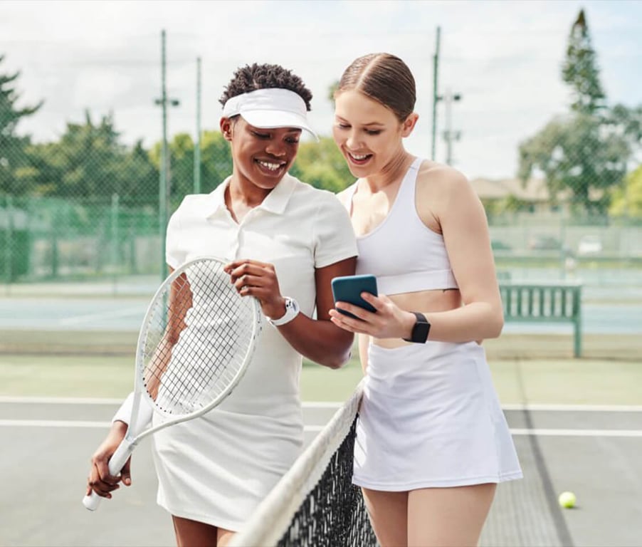 Two women on a tennis court sharing a smartphone, highlighting tennis apps for game improvement.