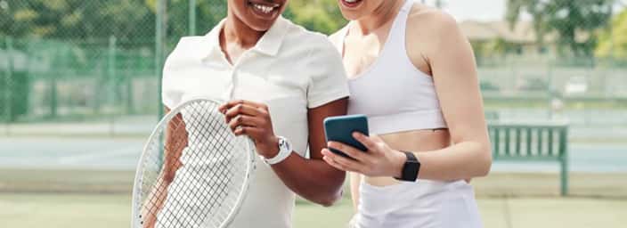 Two women on a tennis court sharing a smartphone, highlighting tennis apps for game improvement.