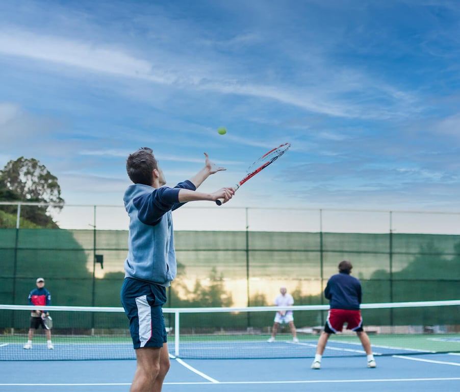 Players engaged in a doubles tennis match, with one player serving the ball on an outdoor court under a clear blue sky.
