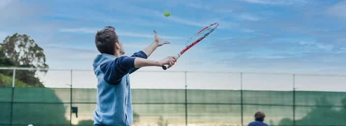 Players engaged in a doubles tennis match, with one player serving the ball on an outdoor court under a clear blue sky.