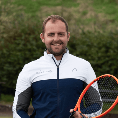 George Cann in a white and blue jacket holding a tennis racket on a tennis court, representing Active Away.