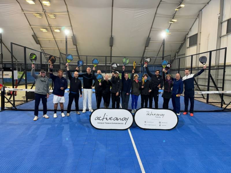 Group of people holding rackets on an indoor tennis court, gathered behind Active Away banners.