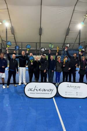 Group of people holding rackets on an indoor tennis court, gathered behind Active Away banners.