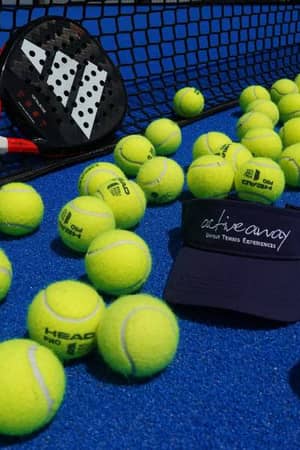 Padel racket and scattered tennis balls on a blue court with an Active Away visor cap nearby.