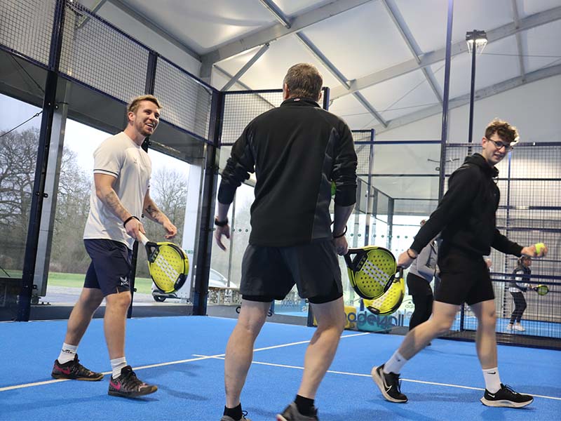 Three men playing padel tennis indoors at Great Western Golf, holding paddles on a blue court.
