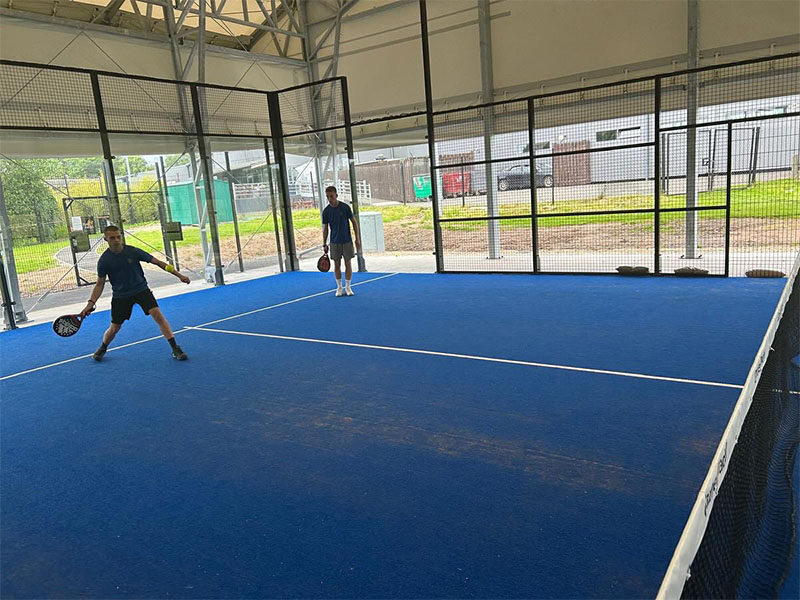 Two men playing padel tennis on a blue indoor court at Great Western Golf facility, with fencing and natural lighting.