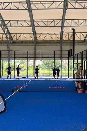 Indoor padel court at Great Western Golf with players in a training session, featuring a blue court, glass walls, and coaching equipment.