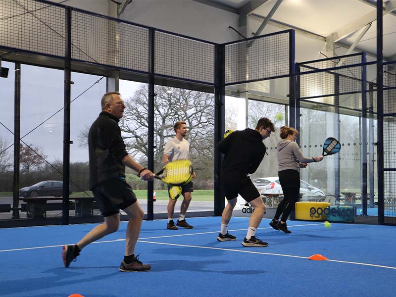 Four people playing padel indoors at Great Western Golf, holding rackets on a blue court with glass walls.
