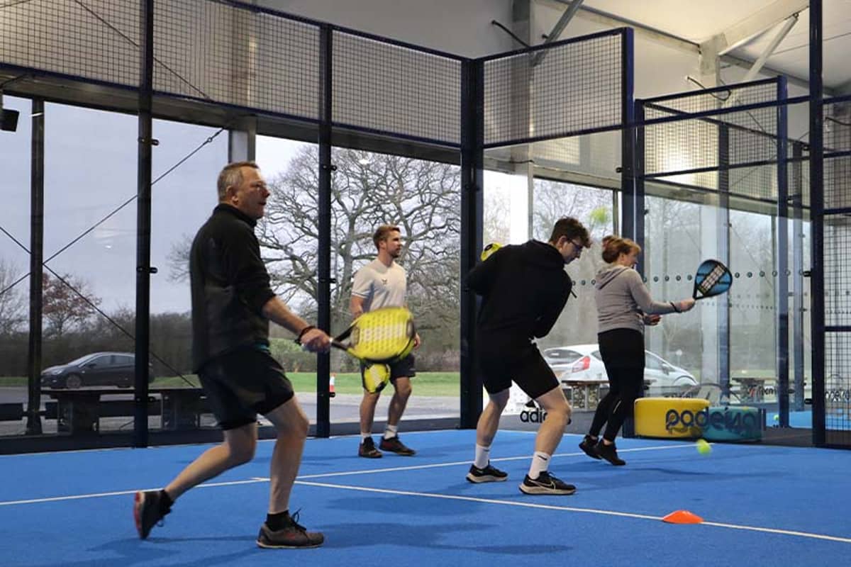 Four people playing padel indoors at Great Western Golf, holding rackets on a blue court with glass walls.