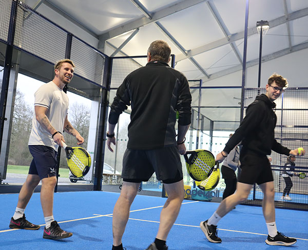 Three men participating in a padel clinic at Great Western Golf on a blue court, holding paddles and wearing sports gear.