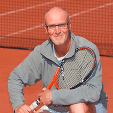 Greg Whitecross on a clay tennis court, smiling and holding a tennis racket, with the net visible in the background.