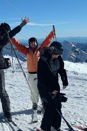Group of six people in ski gear standing on a snowy mountain, with a lodge and mountains in the background, arms raised in celebration.