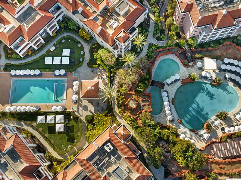 Aerial view of Hilton Vilamoura with two swimming pools and surrounding gardens, featuring sun loungers and red-tiled buildings.