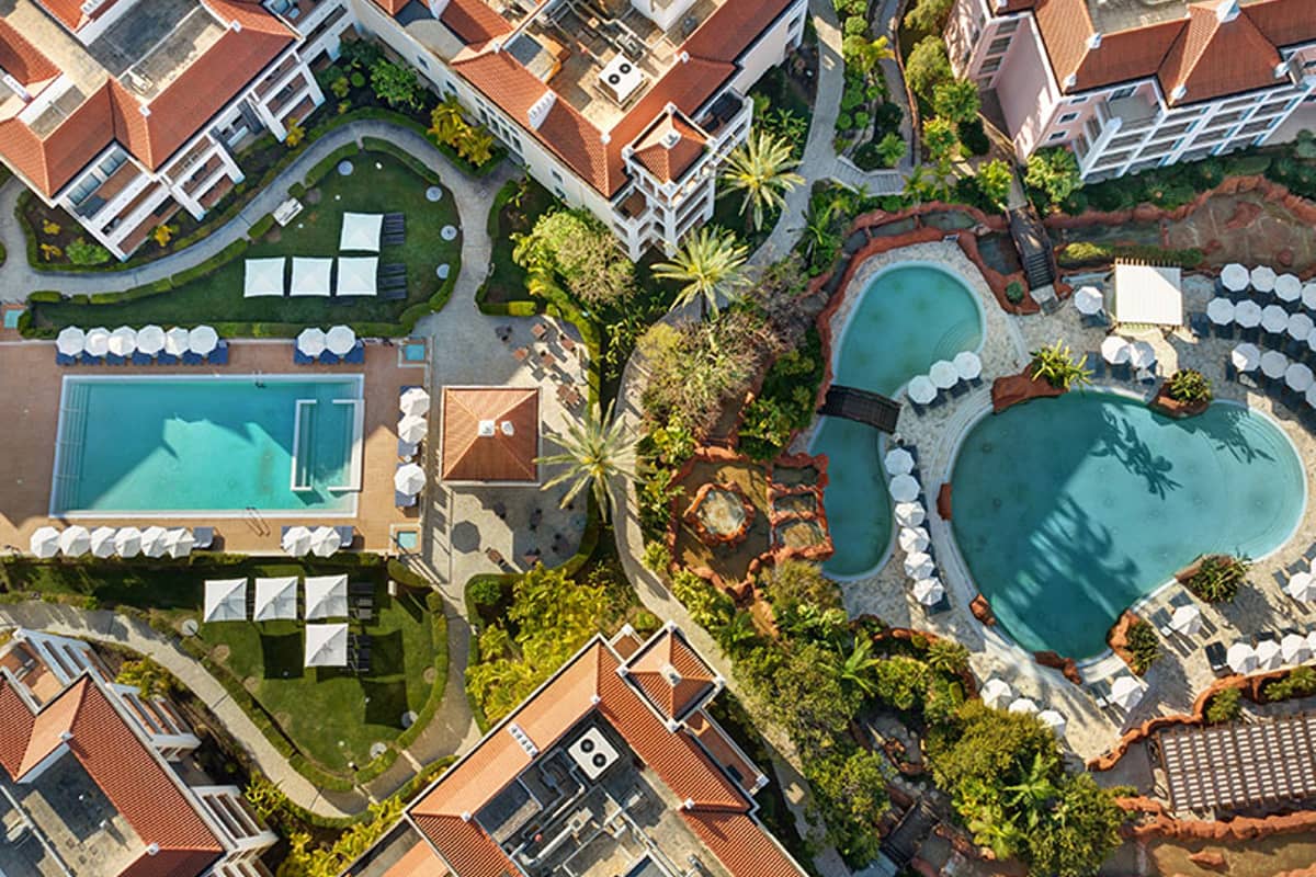 Aerial view of Hilton Vilamoura with two swimming pools and surrounding gardens, featuring sun loungers and red-tiled buildings.