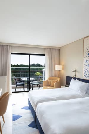 Twin-bedroom suite in Hilton Vilamoura showcasing two beds, abstract art, a desk, flat-screen TV, and a balcony with a green view.