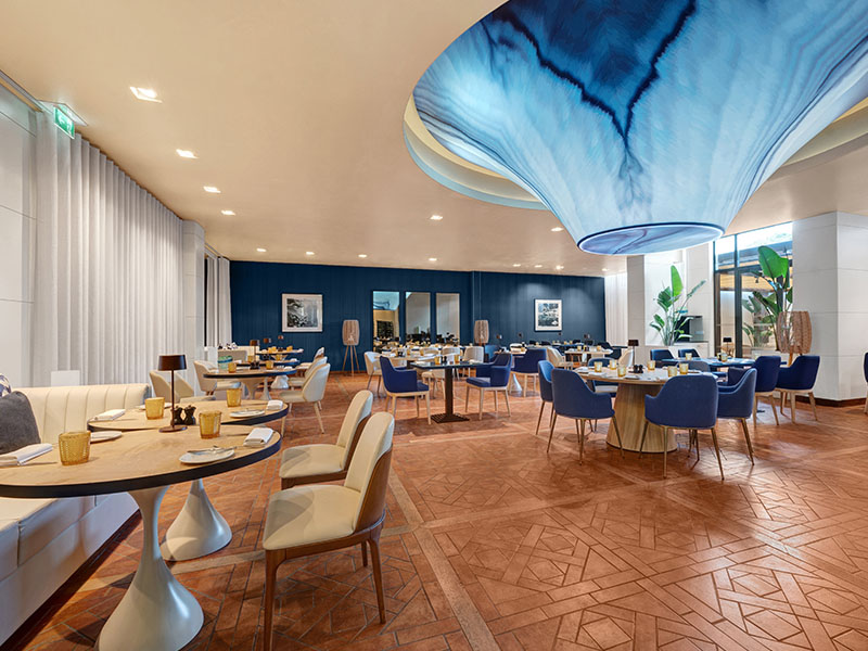 Stylish dining area at Hilton Vilamoura with modern tables, a blue ceiling feature, geometric patterned floor, and natural lighting.