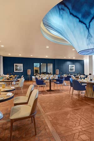 Stylish dining area at Hilton Vilamoura with modern tables, a blue ceiling feature, geometric patterned floor, and natural lighting.