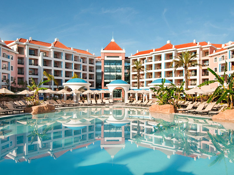 Exterior view of Hilton Vilamoura resort in Portugal, showcasing the central pool, sun loungers, tropical plants, and grand architectural design.