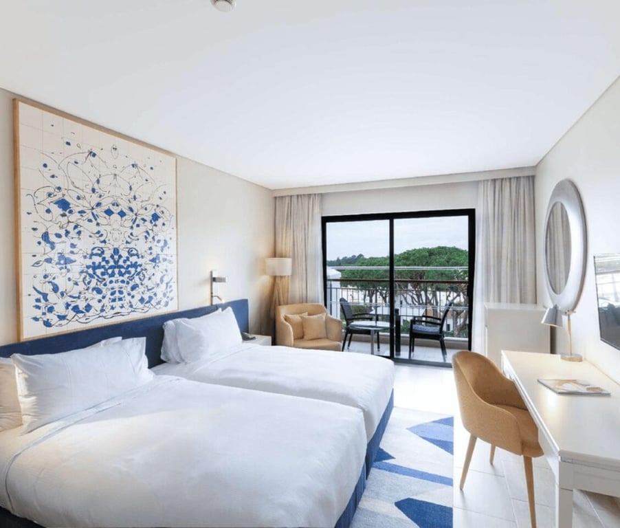 Hilton Vilamoura Deluxe Room with twin beds, a modern abstract painting, a desk with a mirror, and a view of greenery from the balcony.
