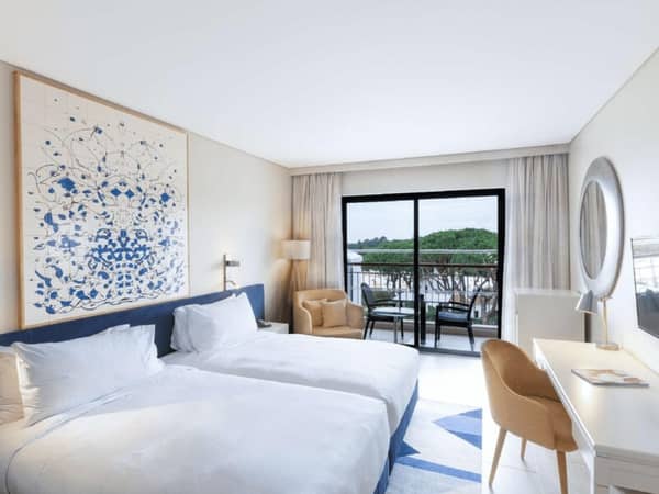 Hilton Vilamoura Deluxe Room with twin beds, a modern abstract painting, a desk with a mirror, and a view of greenery from the balcony.