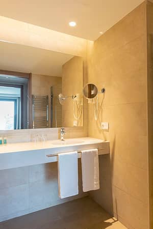 Modern bathroom at Hipotels Barrosa Park with wide vanity, illuminated mirror, sink with toiletries, and a spacious shower area.