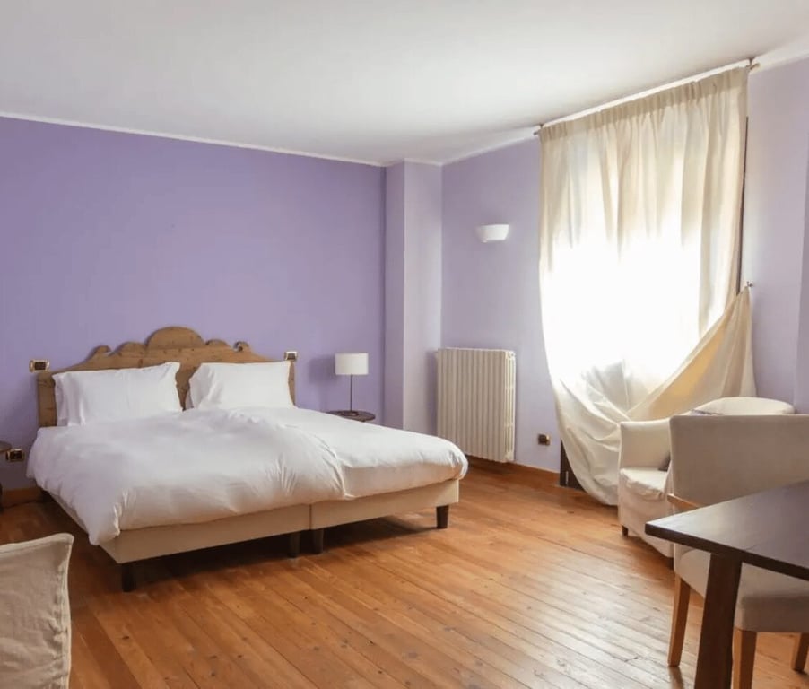 Charme Room at Hotel Cristallo with a double bed, purple walls, wooden flooring, and a cosy seating area beside a large window with curtains.