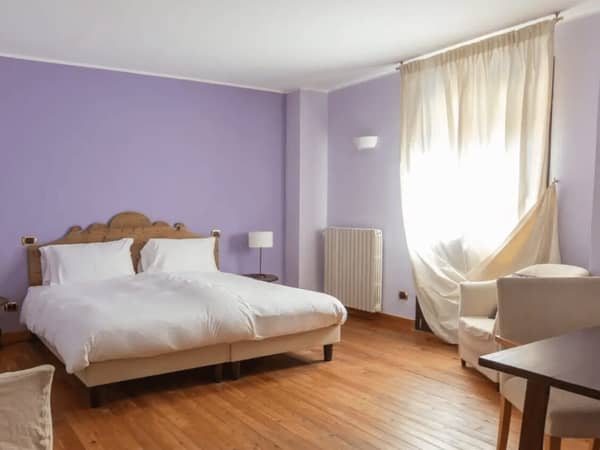 Charme Room at Hotel Cristallo with a double bed, purple walls, wooden flooring, and a cosy seating area beside a large window with curtains.