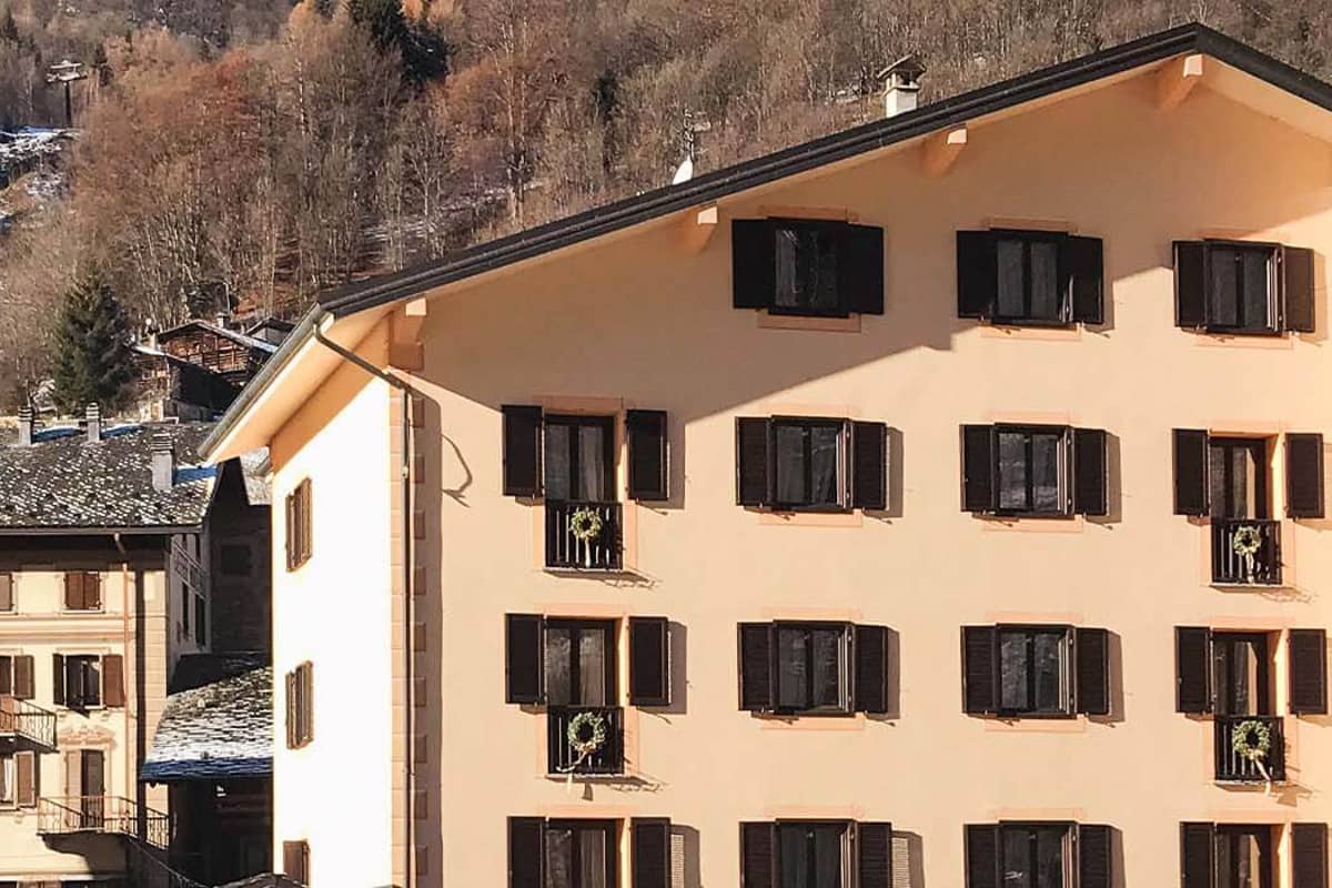 Exterior of Hotel Cristallo with beige facade and dark brown shutters, set against a mountainous backdrop.