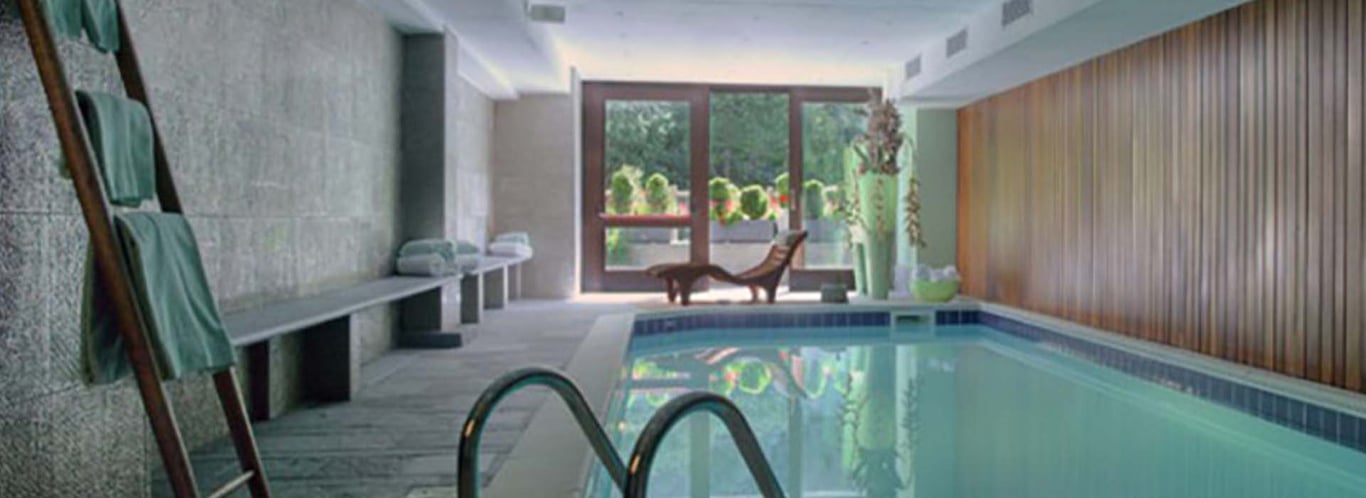Indoor swimming pool at Hotel Cristallo with wooden panelling, folded towels, and a sun lounger by large windows.