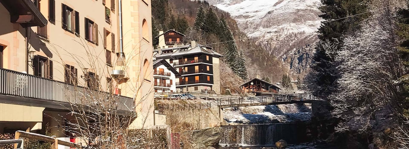 Hotel Cristallo in the Alps, surrounded by snow-laden trees, mountain peaks, and a flowing stream under a clear sky.