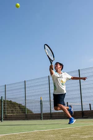 Young tennis player serving on an outdoor court, wearing white shirt and dark shorts, with a clear blue sky in the background.